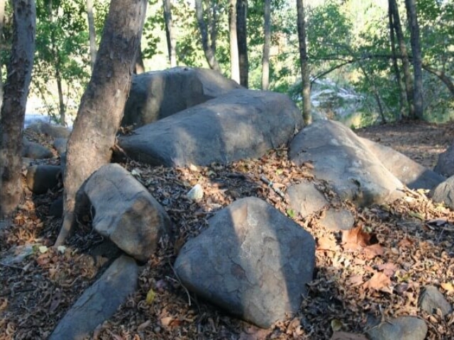 a boulder pile in the foreground with trees on the left and the river peeking through the trees in the background