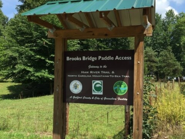 photo of the welcome kiosk at Brooks Bridge Paddle Access a wooden structure with a green peaked roof and a sign, trees in the background
