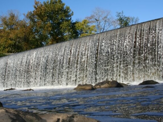 photo of the Haw River rushing over the Glencoe Dam on a sunny day with blue sky and trees in the background and river with rocks in foreground