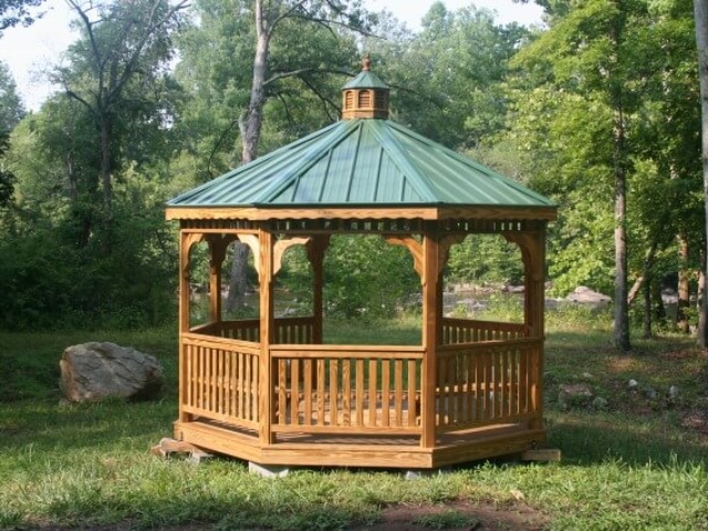 photo of an octagonal wooden gazebo with a green roof in a field with the river and trees in the background