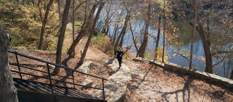 photo of a runner coming up the walkway from the trail to the steps in Saxapahaw with the river in the background