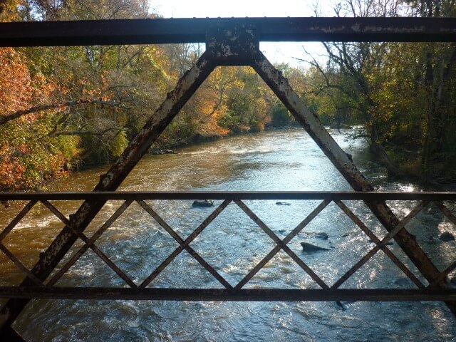 a bird's eye view of the Haw River seen through the metal railings of Goat Island Bridge with trees in Fall color in the background