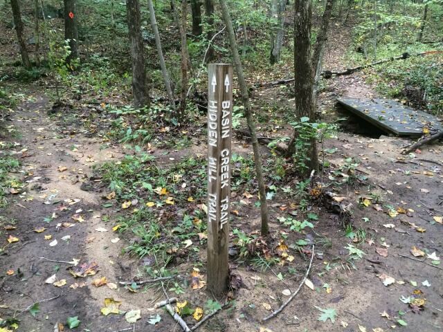 photo of a routed wooden trail marker pointing the way to the Hidden Hill Trail and Basin Creek Trail at a trail crossroads with a footbridge in the background and forest around it all
