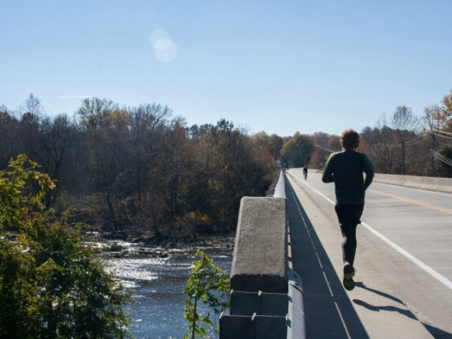 photo of a runner and cyclist on a bridge with the river and trees to the left
