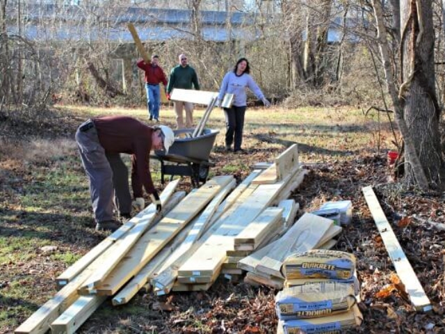 four volunteers carrying wooden boards to a pile of boards with a wheelbarrow and bags of concrete mix in the foreground