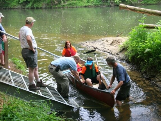 a canoe with a family pulled up to the steps in the river with several people assisting them with getting out of the canoe