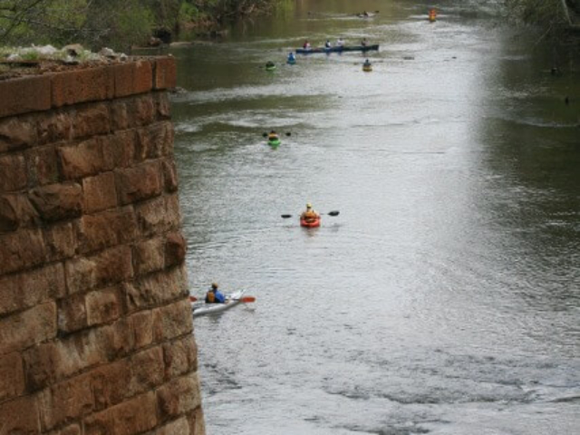 photo of a line of paddlers on the river with a brick bridge piling in the foreground and trees in the background